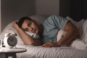 A man lays wide awake in bed, staring at his clock. He is experiencing insomnia and depression.