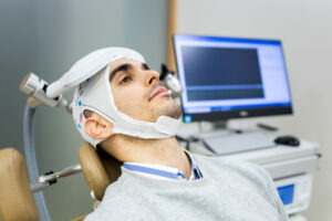 A man is utilizing TMS for treatment-resistant depression. He's in a doctor's office chair with a TMS coil lying gently against his forehead. It's cushioned by a light head wrap that's secured by velcro beneath his chin.
