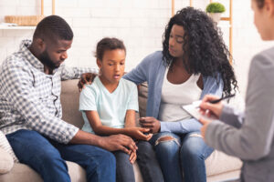 A mom, dad, and daughter sit together during a family couseling session in Annapolis, MD. They each have an arm around their daughter, comforting her.