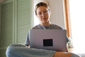 A teen girl sits cross-legged with her laptop. She is participating in online therapy for her depression.