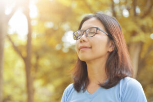 A woman is standing outside in a wooded area. She is closing her eyes and using her senses to practice being present, a mindfulness technique.
