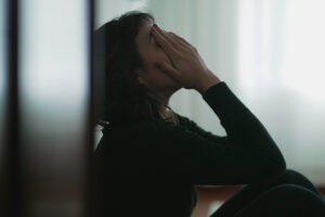 A woman sits with her back to the wall in a dimly lit room. She is covering her face with her hands. She's experiencing symptoms of clinical depression.
