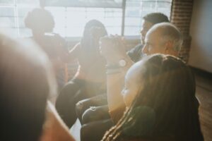 Group of people sitting together in a sunlit room hold hands in support, creating a sense of unity and encouragement.